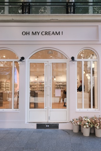 Façade avec portes vitrées, fenêtres cintrées et bouquets de fleurs devant l'entrée dans la boutique Oh My Cream !.
