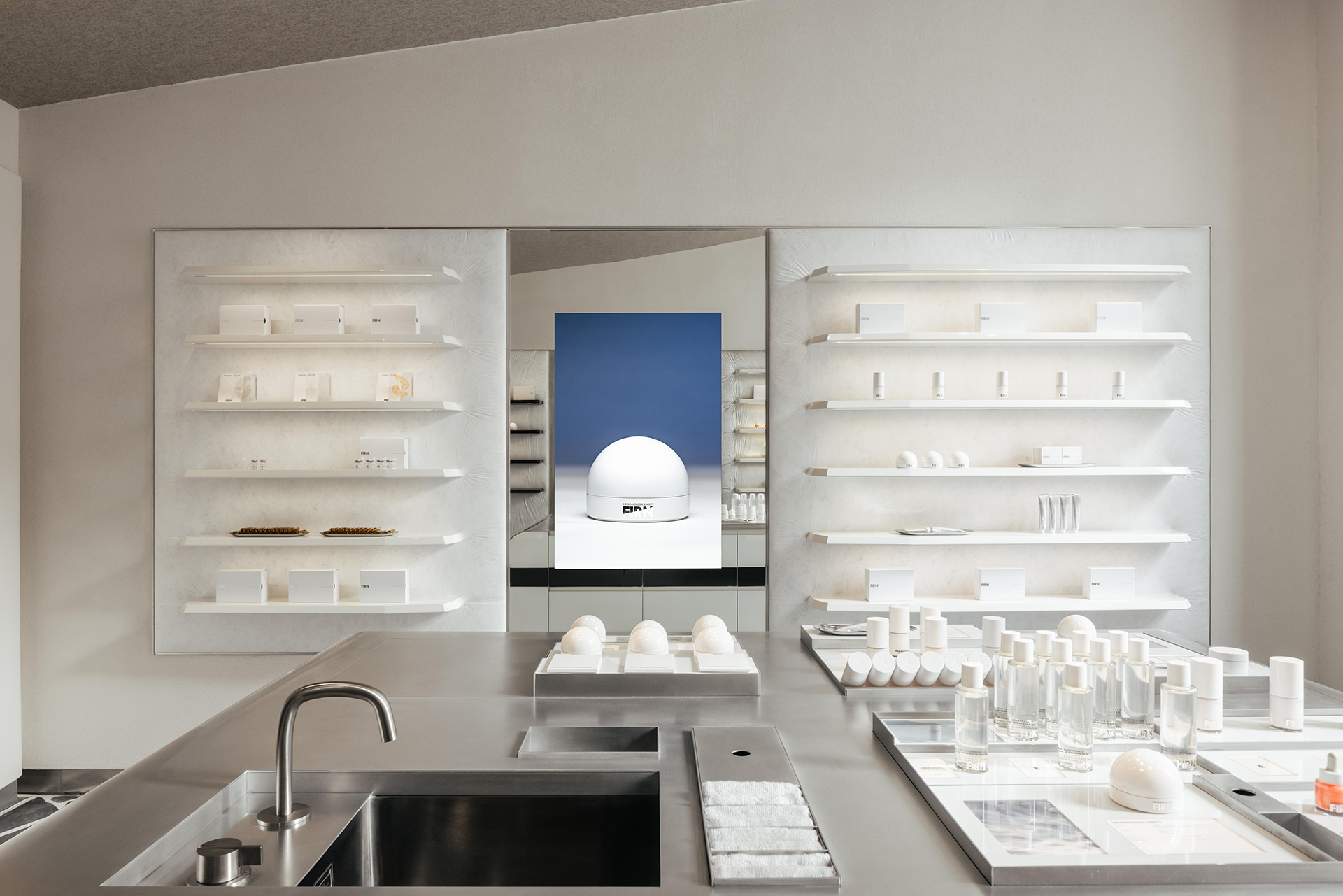 Intérieur épuré avec étagères blanches, présentoirs, lavabo et grand miroir reflétant un mur bleu et une sculpture, projet d'architecture retail de luxe.