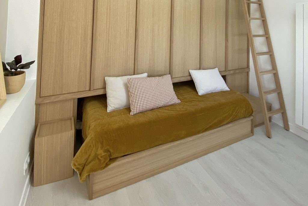 Intérieur clair avec un lit de jour en bois, couverture jaune moutarde et échelle en bois, projet d'architecture retail de luxe.