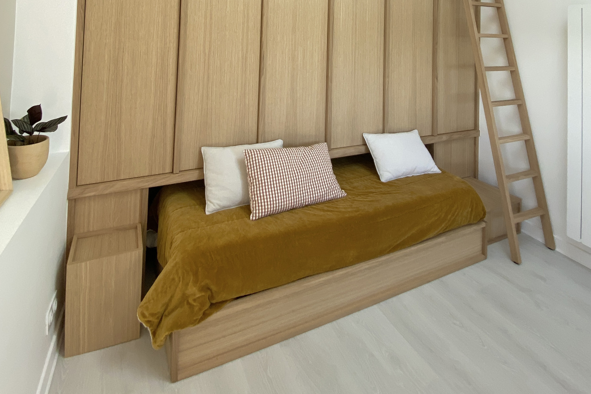 Intérieur clair avec un lit de jour en bois, couverture jaune moutarde et échelle en bois, projet d'architecture retail de luxe.