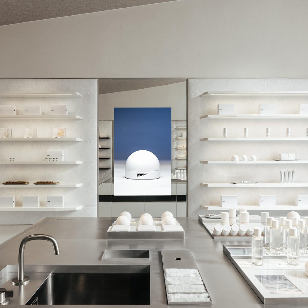Intérieur épuré avec étagères blanches, présentoirs, lavabo et grand miroir reflétant un mur bleu et une sculpture, projet d'architecture retail de luxe.
