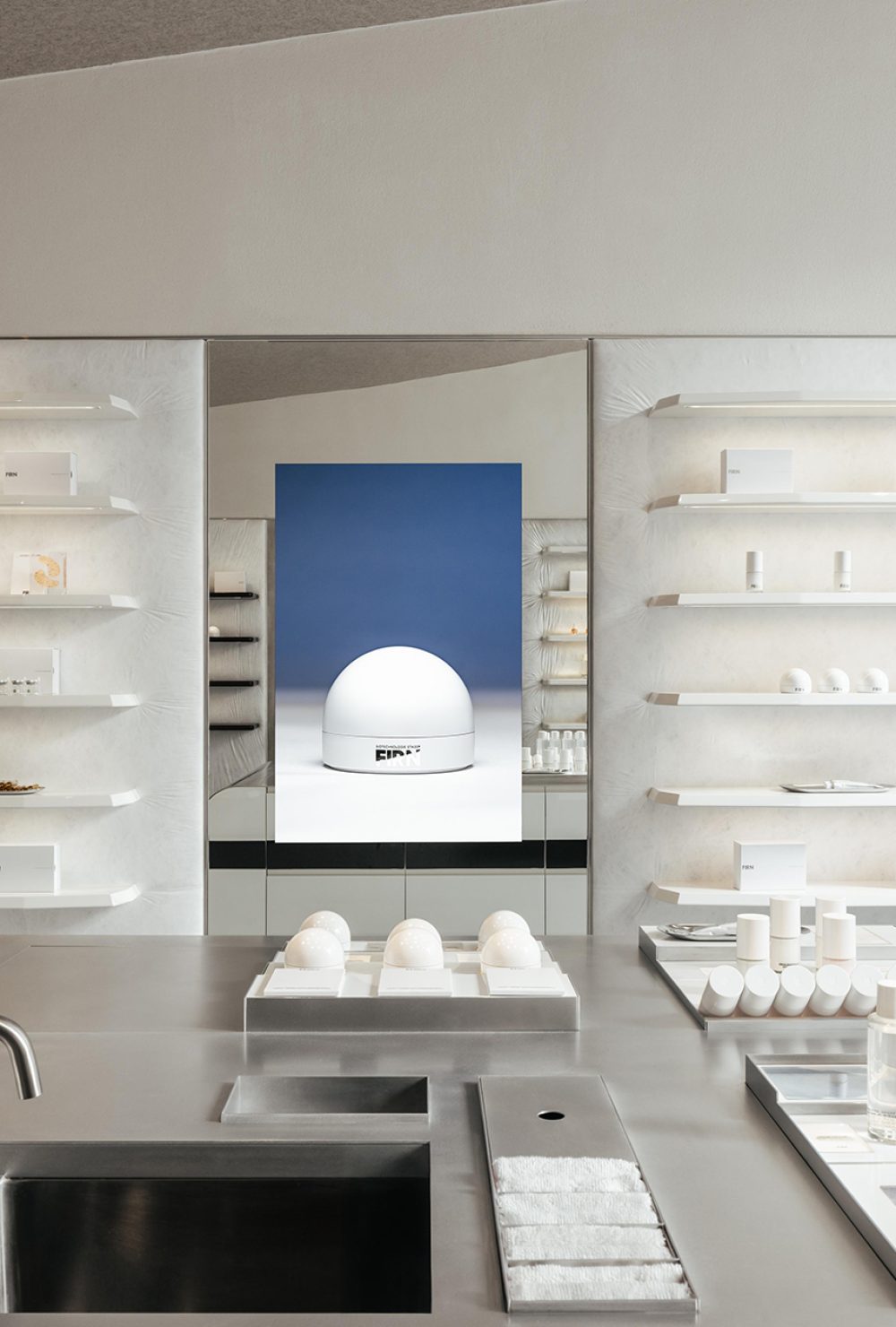 Intérieur épuré avec étagères blanches, présentoirs, lavabo et grand miroir reflétant un mur bleu et une sculpture, projet d'architecture retail de luxe.