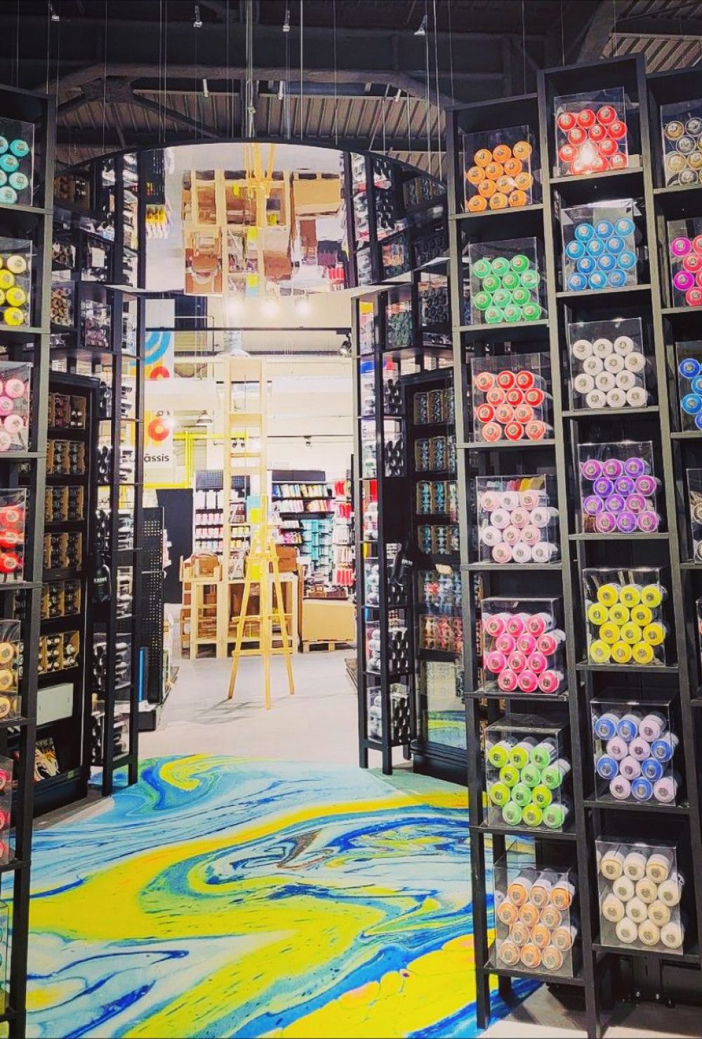 Etagères avec des bombes de peinture colorées et un sol marbré de peinture bleu et jaune dans la boutique Rougier&Plé de Claye-Souilly.